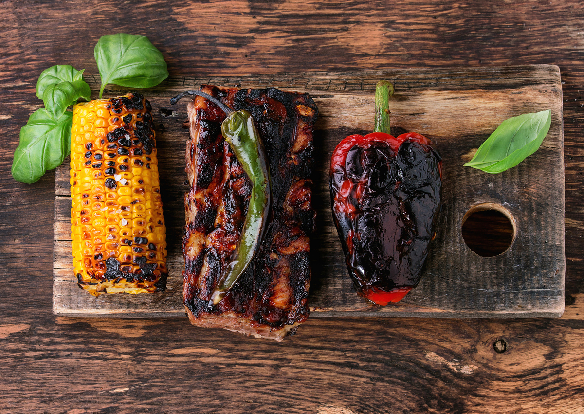 BBQ grilled pork ribs, corn, green chili and a red bell pepper are served on wooden chopping board.