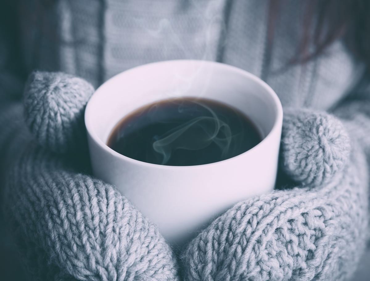 A person wearing mittens holds a cup of steaming black coffee.