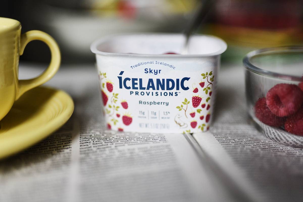 An empty yogurt container sits on a table with a mug and raspberries.